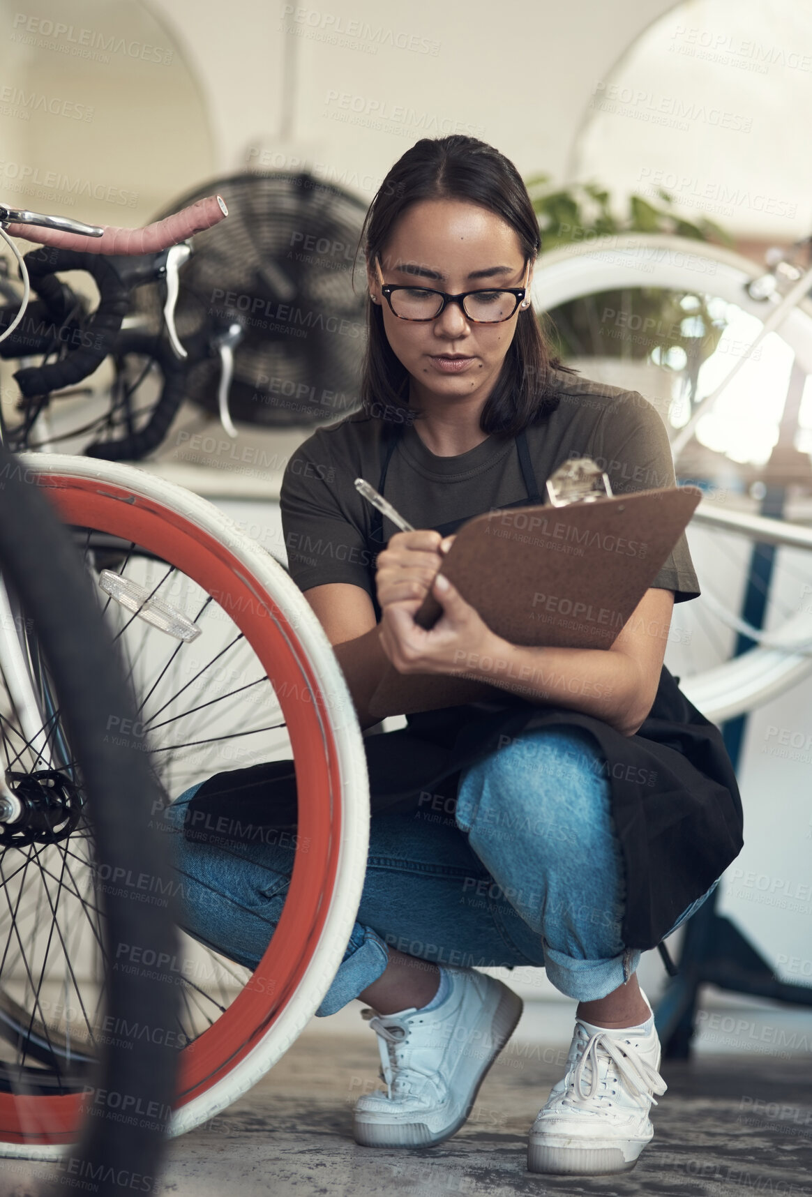 Buy stock photo Full length shot of an attractive young woman crouching in her bicycle shop and holding a clipboard