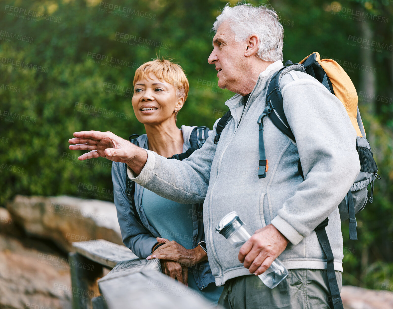 Buy stock photo Couple, elderly and hiking in forest with fitness and interracial, talking about view and relax while trekking. Retirement, exercise and hike in nature with water bottle for hydration and travel.