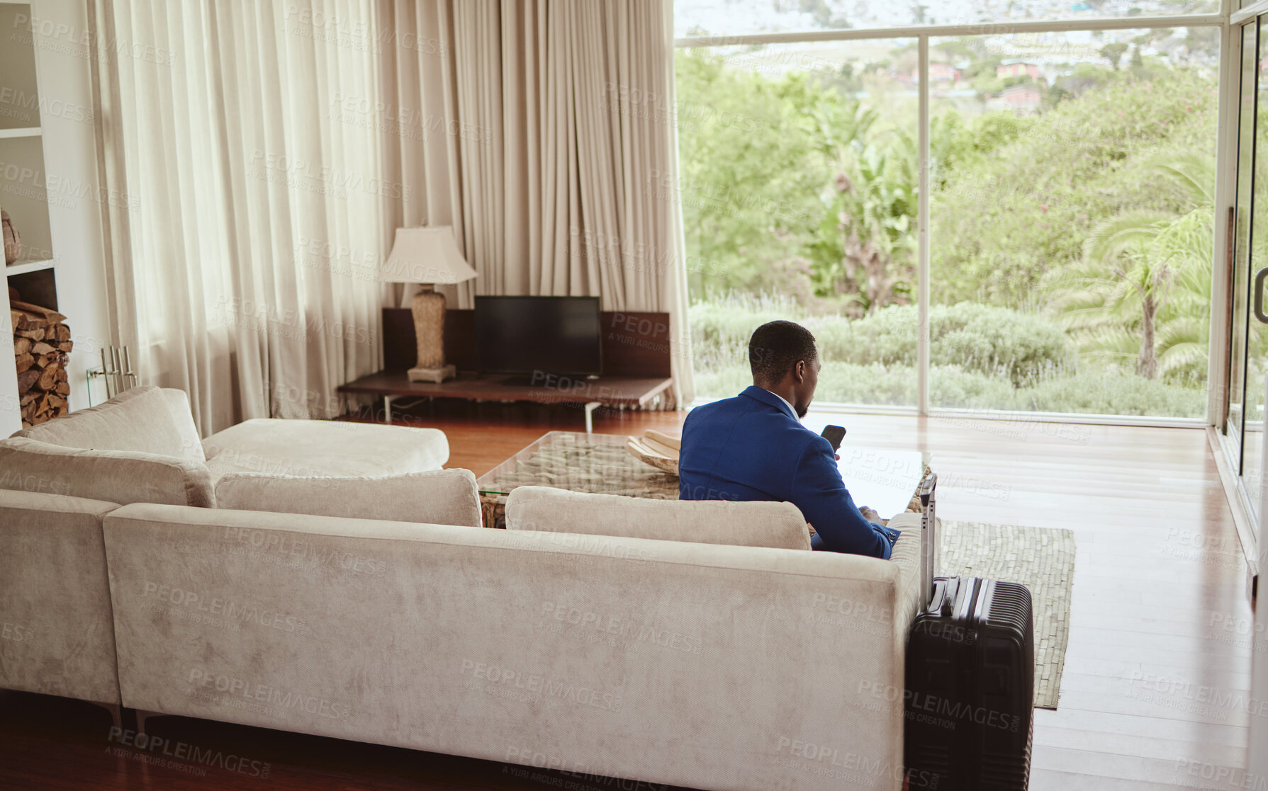 Buy stock photo Phone, travel and hospitality with a business black man sitting on a sofa in the living room of a hotel with his luggage. Back, bag and communication with a male employee typing a text message