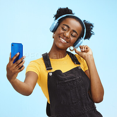 Buy stock photo Selfie, smartphone and black woman with headphones, social media and connection on blue studio background. African American female influencer, girl and content creator with smile and online streaming