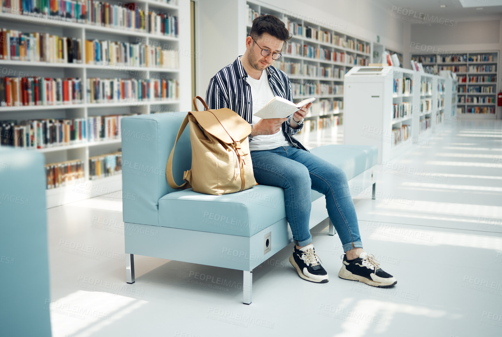 Buy stock photo Library, reading and man with book on sofa for education, studying and academic research. University, scholarship and male student on couch for learning, hobby and knowledge in college bookstore
