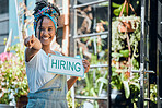 Small business, black woman or business owner with a hiring sign at door entrance in floral retail store. Advertising, marketing and happy entrepreneur pointing with job offer message at flower shop