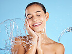 Water splash, beauty and woman isolated on blue background for skincare, cosmetics cleaning and happy face. Smile of person or model washing facial for dermatology, wellness and aesthetic in studio
