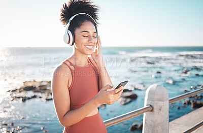 Buy stock photo Black woman at beach, headphone and smartphone with fitness, runner listening to music for sports motivation. Happy, streaming online with podcast or radio, phone and running by sea with calm outdoor