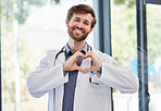 Doctor, man and heart hands with smile at hospital for support, cardiology or kindness by window. Medic portrait, hand sign or medical service with icon of care, love or help for consulting at clinic