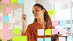 Focused female designer planning ideas on a glass wall with colorful sticky notes inside a modern and creative office. Busy woman enjoying her job while brainstorming projects and managing projects