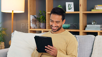 Young man browsing on a digital tablet on a sofa at home. Happy guy smiling and laughing while watching funny videos, scrolling on social media and streaming movies online to enjoy over the weekend