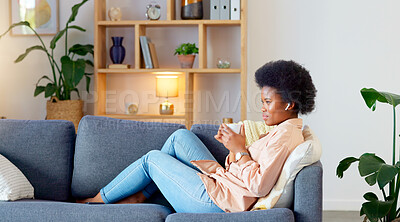 Relaxed Black woman listening to a podcast while drinking coffee and sitting on a couch at home. Happy African American woman streaming online content, enjoying an audiobook while laughing