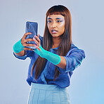 Fashion, selfie and woman with smartphone and cyberpunk neon clothing isolated on blue background. Social media, future and trendy gen z influencer from India with phone in studio for profile picture
