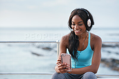 Buy stock photo Black woman, phone and headphones at beach, sitting and smile for video, music or funny podcast to relax. Runner, rest and smartphone with meme, social media app with happiness on outdoor adventure