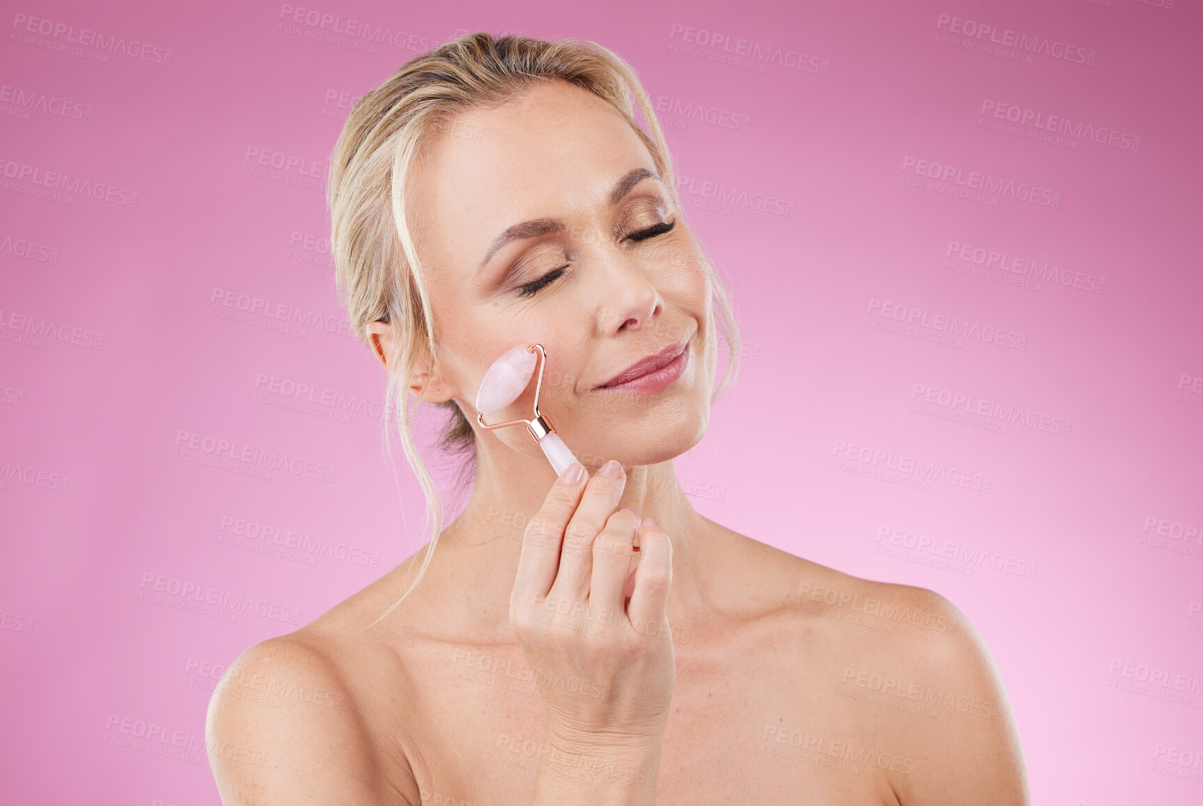 Buy stock photo Beauty, facial roller and woman doing face massage with dermatology and cosmetics in studio. Mature aesthetic person on pink background for skincare, self care and rose quartz results for skin glow