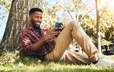 Buy stock photo Black man, phone and relaxing by tree in a park for social media, communication or chatting lying on green grass. Happy African American male with smile on smartphone for 5G connection in nature