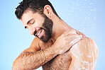 Shower, cleaning and man with soap, smile and water splash in studio for wellness, hygiene and grooming. Skincare, healthy skin and male with foam, cosmetics and washing body on blue background 