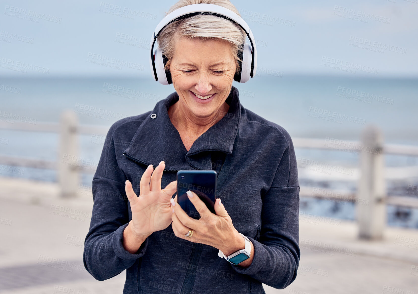 Buy stock photo Senior woman, phone and headphones listening to music by the beach with smile for audio track. Happy elderly female smiling with smartphone for online streaming or 5G connection by the ocean coast