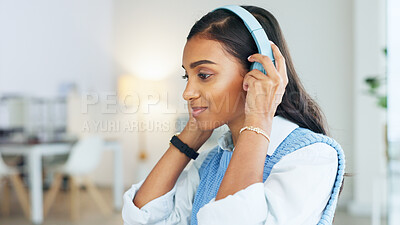 Side view of young Indian woman sitting in office with blurred background. Millenial worker putting headphones on and smiling while moving to the music. Intern looking confident at her new job