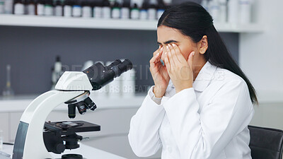 Female scientist looking through microscope and rubbing her eyes while suffering from eye strain or discomfort. Stressed biotechnology specialist does analysis of test sample in medical lab