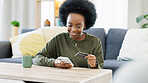 Young black woman shopping online using her phone and bank credit card in a bright living room. Banking and managing assets and finance using convenient wireless technology from the comfort of home