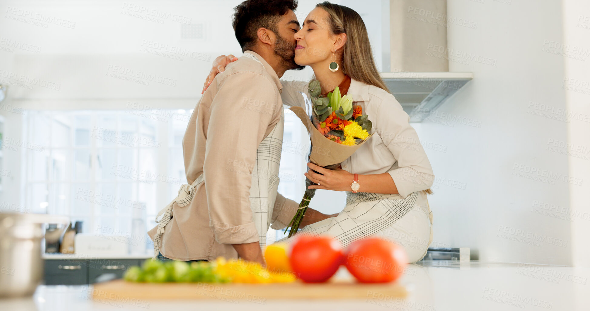 Buy stock photo Couple, cooking and flowers for gift in kitchen, hug and smile with romance, love and care with kiss. Man, woman and celebrate with food together, embrace and floral bouquet for diet with nutrition