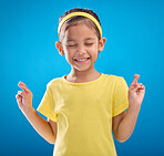 Child, hand and fingers crossed in studio for hope, wish or good luck against a blue background. Girl, eyes closed and emoji hands, praying and wishing, happy and smile while posing on isolated space