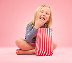 Popcorn eating, portrait and happy girl in a studio with pink background sitting with movie snacks. Food taste, happiness and hungry young child with a paper bag and chips feeling relax with a smile