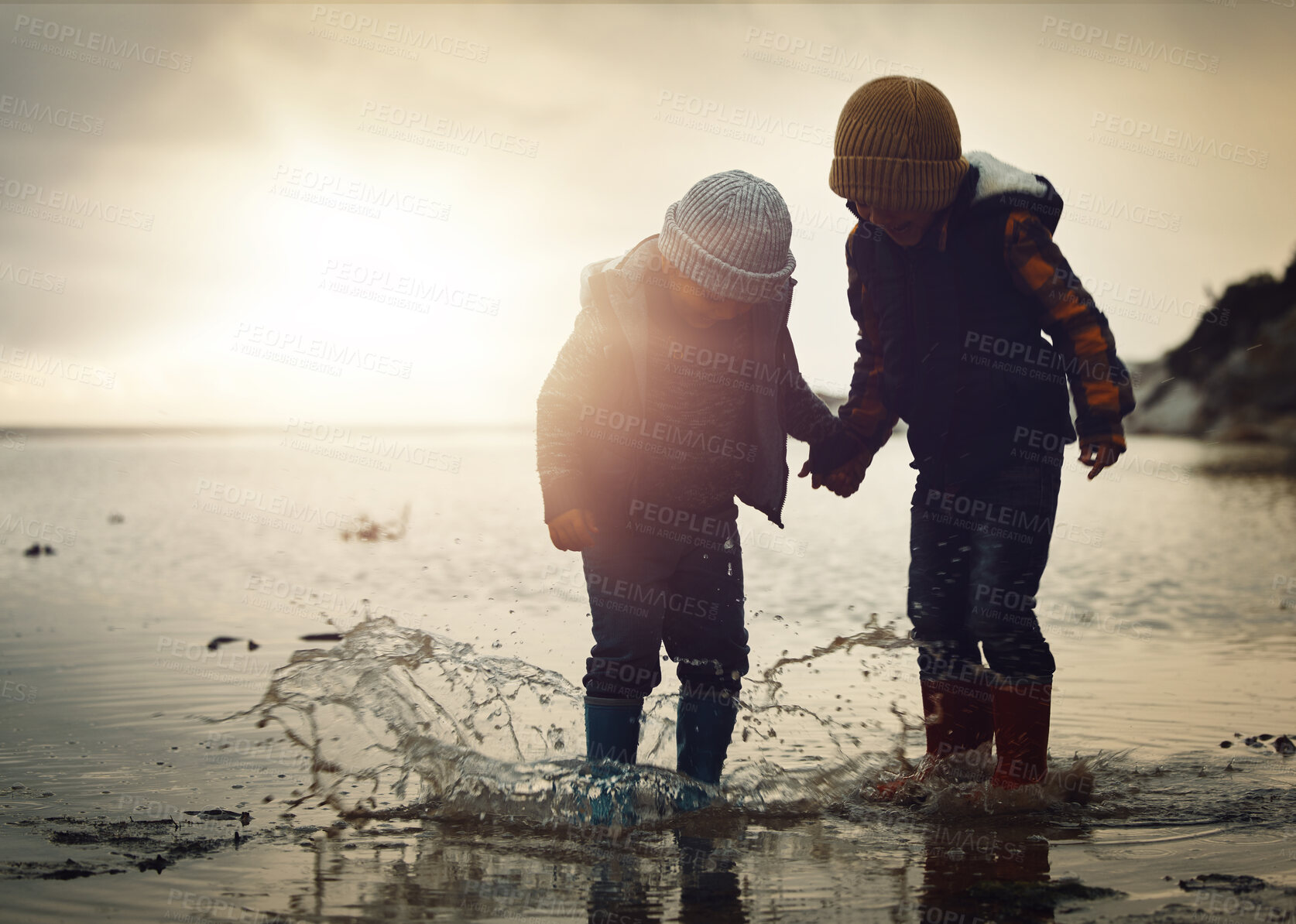 Buy stock photo Beach, sunset and silhouette of children in water splash together, holding hands and playing in waves. Fun, holiday and brothers, happy in boots on ocean vacation, jumping in sea on winter evening.