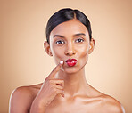 Beauty, red lipstick and makeup portrait of a woman in studio for cosmetics, skin glow and shine. Indian female model with finger on face or kiss on beige background for self care and lips with color