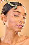 Face, skincare roller and woman with eyes closed in studio isolated on a brown background. Dermatology, facial massage and Indian female model with jade crystal for healthy skin treatment and beauty.