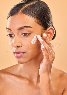 Buy stock photo Face, beauty skincare and woman with cream in studio isolated on a brown background. Dermatology idea, cosmetics and thinking Indian female model apply lotion, creme and moisturizer for healthy skin.