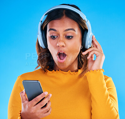 Buy stock photo Headphones, cellphone and shocked woman in a studio listening to a podcast, music or radio. Surprise, technology and female model streaming an album, song or playlist with a phone by blue background.