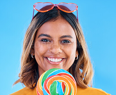 Buy stock photo Portrait, lollipop and a woman smiling on a blue background in studio wearing heart glasses for fashion. Face, candy and sweet with a happy young female eating a giant snack while feeling positive