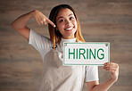 Happy woman, portrait and pointing to hiring sign for small business recruitment, career or job opportunity against studio background. Female entrepreneur with apron holding billboard poster for hire