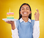 Smile, wish and woman with cake in studio for happy celebration or party on yellow background. Happiness, excited gen z model with fingers crossed and candle in rainbow dessert to celebrate milestone