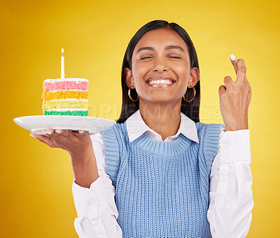 Buy stock photo Smile, wish and woman with cake in studio for happy celebration or party on yellow background. Happiness, excited gen z model with fingers crossed and candle in rainbow dessert to celebrate milestone