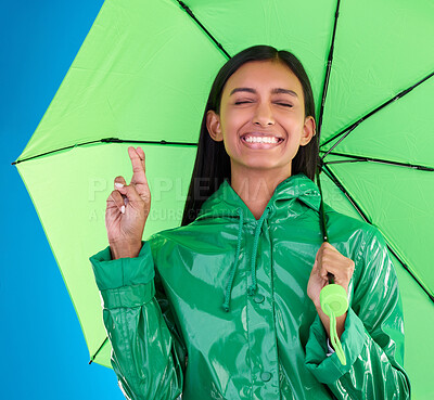 Buy stock photo Green, umbrella and fingers crossed with a woman in studio on a blue background during winter for insurance. Rain, fashion or cover and an attractive young female wishing for weather change with hope