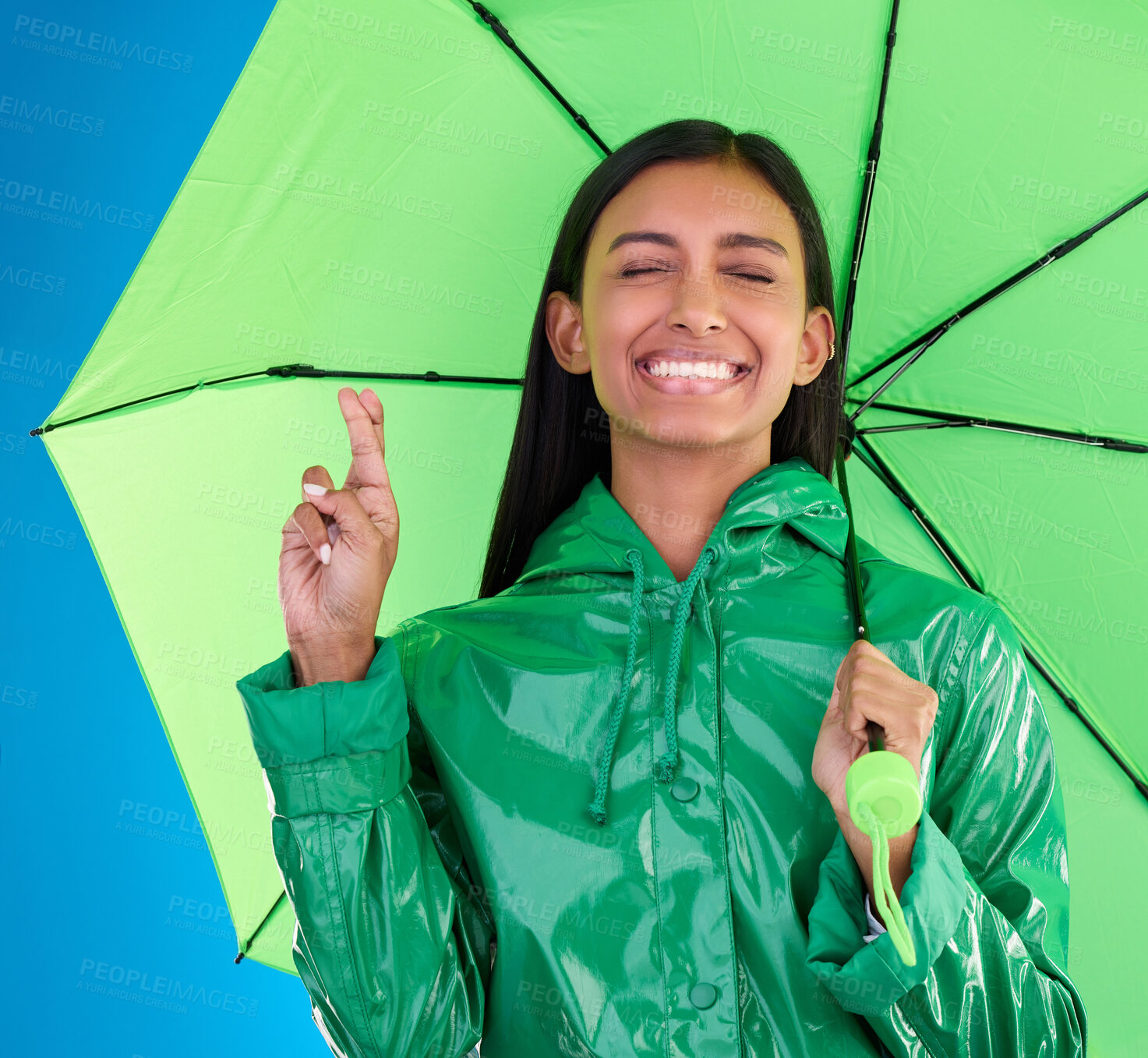 Buy stock photo Green, umbrella and fingers crossed with a woman in studio on a blue background during winter for insurance. Rain, fashion or cover and an attractive young female wishing for weather change with hope