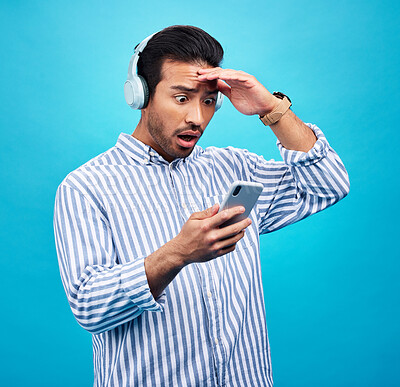 Buy stock photo Phone, headphones and shock with a man on a blue background in studio reading news on social media. Wow, surprise or podcast with a male listening to audio in disbelief at hearing mind blowing gossip