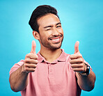 Portrait, thumbs up and wink by asian man in studio with positive, feedback or review on blue background. Face, smile and hand gesture by male showing yes, agreement and vote emoji while isolated