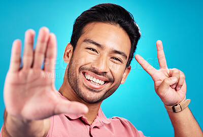 Buy stock photo Happy asian man, portrait and peace sign for selfie, profile picture, or social media against a blue studio background. Male influencer or vlogger with smile showing peaceful emoji for photo or vlog
