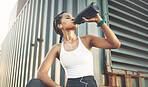 One fit young hispanic woman wearing earphones and taking a rest break to drink water from a bottle while exercising outdoors. Female athlete quenching thirst and cooling down after run and training workout in an urban setting