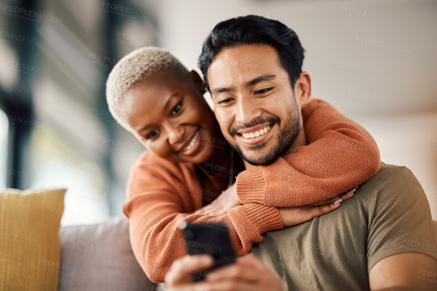 Buy stock photo Phone, interracial couple and technology on a sofa at home reading text in a living room. Support, young people and hug together in a house sitting on a lounge couch with mobile streaming and video