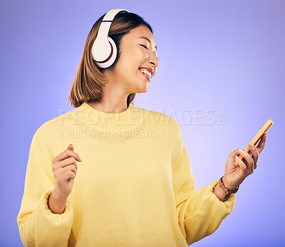 Buy stock photo Music, phone and happy Asian woman in studio for social media, internet meme and online chat. Headphones, smile and female person on smartphone listening to audio, song and track on purple background