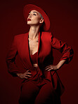 Confident, fashion and a model woman on a red studio background for elegant or trendy style. Aesthetic, art and beauty with a young female person standing hands on hips in an edgy or classy suit