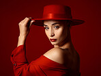 Portrait, fashion and a confident woman on a red studio background for elegant or trendy style. Face, aesthetic and hat with a young female model in an edgy or classy outfit for aesthetic beauty