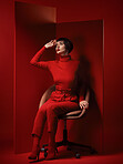 Thinking, fashion and a model woman on red background in studio for elegant, chic or trendy style. Aesthetic, art and beauty with an edgy or classy young person in unique clothes or suit on a chair