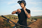 Sports, bike and man checking his pulse on a mountain dirt road in nature for race or competition training. Fitness, exercise and young male biker athlete with watch for the time at an outdoor trail.