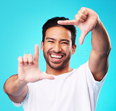 Buy stock photo Happy, hand frame and portrait of a man for a selfie, creative aesthetic or advertising photography. Smile, laughing and face of an Asian person with a gesture for a photo on a blue background