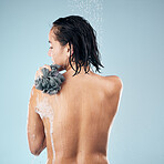 Woman, shower and back in water drops with loofah in hygiene, grooming or washing against a blue studio background. Rear view of female person in body wash, cleaning or skincare with rain in bathroom