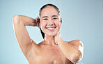 Face, shower and happy woman washing hair in studio isolated on blue background. Water splash, hygiene and portrait of natural Asian model smile, cleaning or bathing for wellness, beauty and skincare