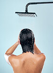 Woman, shower and back in water drops for hygiene, grooming or washing against a blue studio background. Rear view of female person in body wash, cleaning or skincare routine under rain in bathroom
