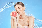 Woman, splash and fruit with water for shower, skincare and bodycare for health and wellness on a blue studio background. Beauty, wash and hydration with grapefruit or vitamin c for cosmetic cleanse